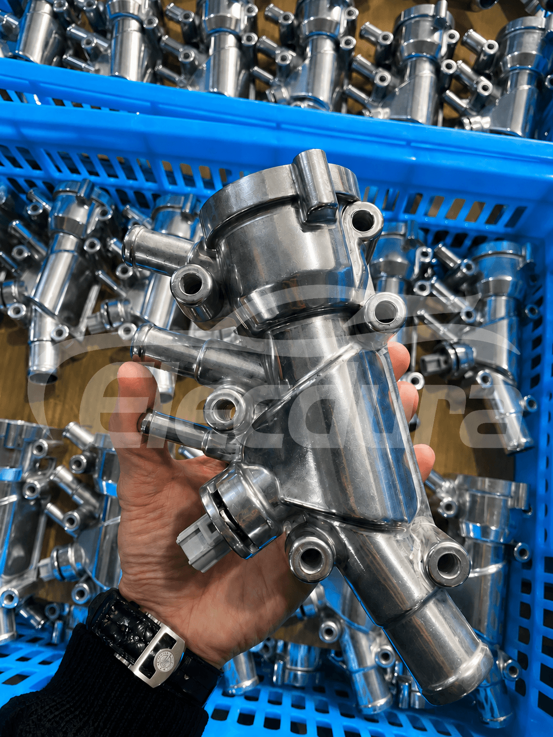 Aluminum thermostat housing assembly held in front of bulk engine cooling parts in blue factory bins.png